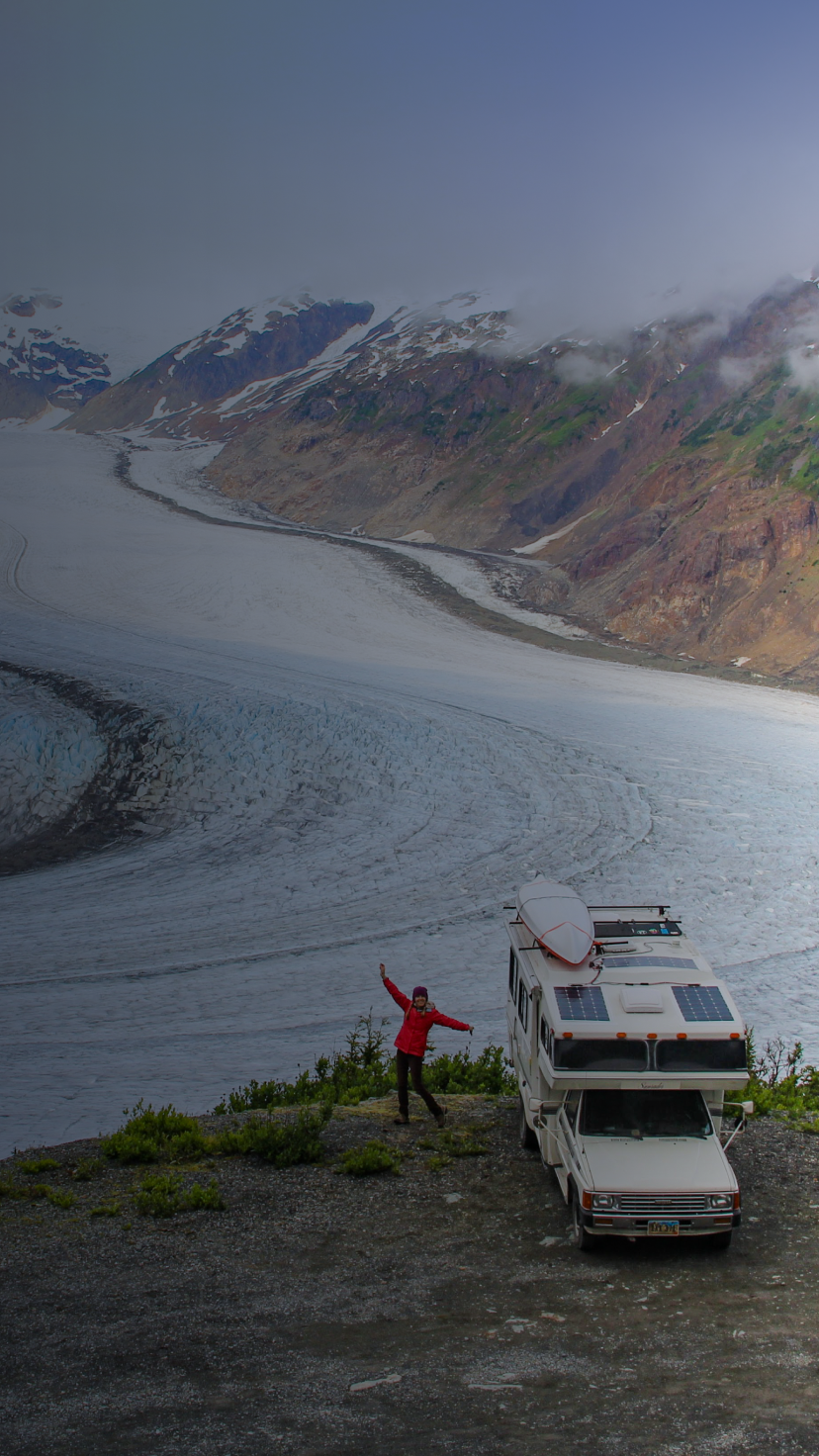 A woman posing next to an RV on a cliff with a snowy glacier below. 